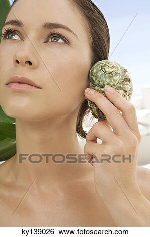 Stock Photograph - Portrait of a young woman holding a shell against her ear, outdoors. Fotosearch