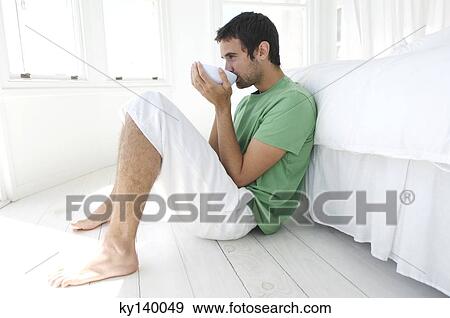 Stock Photo - Young man drinking coffee, leaning against bed. Fotosearch