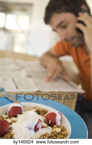Young man reading newspaper, using mobile phone, strawberries and cereals in foreground View Large Photo Image Picture - Young man reading newspaper, using mobile phone, strawberries and cereals in foreground. Fotosearch