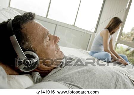 Man lying on bed, listening to music with headphones, woman in background View Large Photo Image Stock Photograph - Man lying on bed, listening to music with headphones, woman in background. Fotosearch