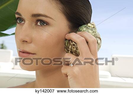 Stock Photo - Portrait of a young woman holding shell against her ear, outdoors. Fotosearch