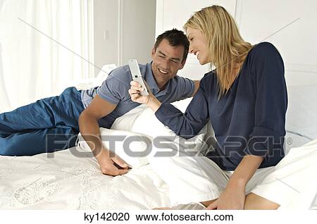 Stock Image - Young couple in bed, using mobile phone. Fotosearch