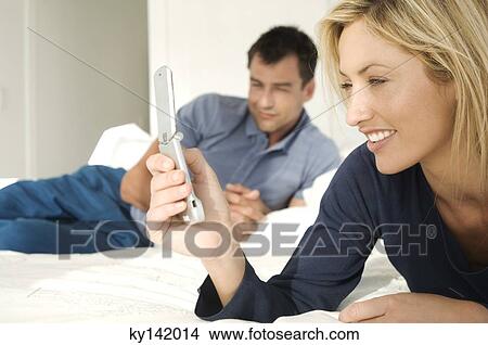 Young couple in bed, woman using mobile phone, man in background View Large Photo Image Picture - Young couple in bed, woman using mobile phone, man in background. Fotosearch