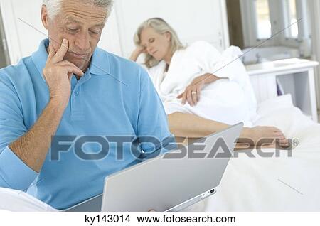 Man using laptop in bedroom, woman in background View Large Photo Image Picture - Man using laptop in bedroom, woman in background. Fotosearch