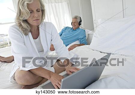 Stock Image - Woman using laptop in bedroom, man with headphones in background. Fotosearch