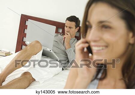 Woman using mobile phone, man on bed using laptop in background View Large Photo Image Stock Photography - Woman using mobile phone, man on bed using laptop in background. Fotosearch