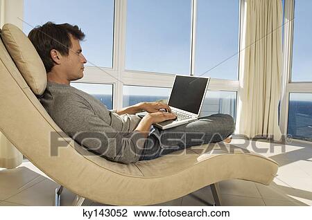Young man using laptop in armchair View Large Photo Image Stock Image - Young man using laptop in armchair. Fotosearch