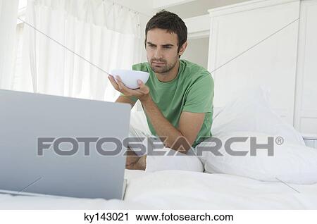 Stock Image - Young man using laptop in bed. Fotosearch