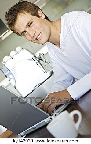 Stock Image - Young man using laptop in kitchen. Fotosearch