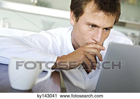 Stock Image - Young man using laptop in kitchen. Fotosearch