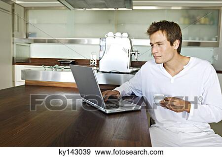 Stock Photo - Young man using laptop in kitchen. Fotosearch
