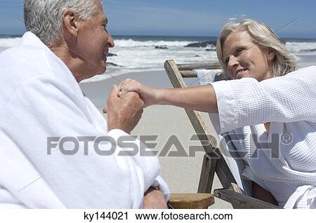 Stock Image - Couple in bathrobe holding hands, sitting on beach chairs. Fotosearch