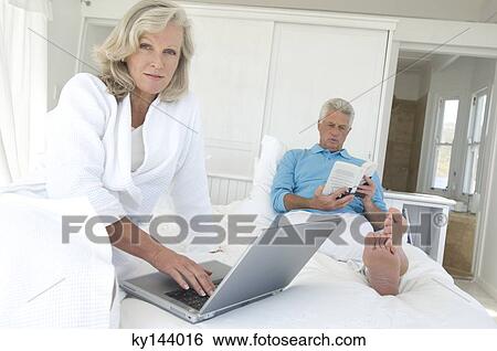 Stock Photograph - Couple in bed, man reading, woman using laptop. Fotosearch