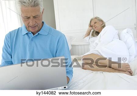 Couple in bedroom, man using laptop, woman lying in background View Large Photo Image Stock Image - Couple in bedroom, man using laptop, woman lying in background. Fotosearch