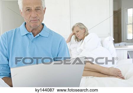 Couple in bedroom, man using laptop, woman lying in background View Large Photo Image Stock Photo - Couple in bedroom, man using laptop, woman lying in background. Fotosearch