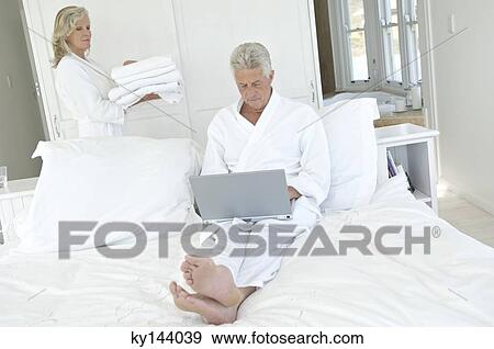 Couple in bedroom, woman carrying stack of towels, man using laptop View Large Photo Image Stock Photo - Couple in bedroom, woman carrying stack of towels, man using laptop. Fotosearch