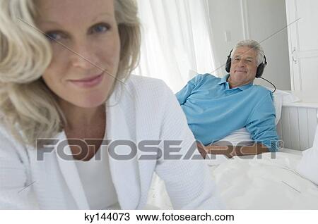 Couple in bedroom, woman looking at camera, man listening to music with headphones in background View Large Photo Image Stock Image - Couple in bedroom, woman looking at camera, man listening to music with headphones in background. Fotosearch