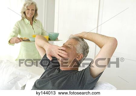 Stock Image - Woman bringing breakfast to man yawning in bed. Fotosearch