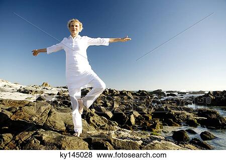 Stock Photo - Senior woman doing yoga on seaside rocks. Fotosearch