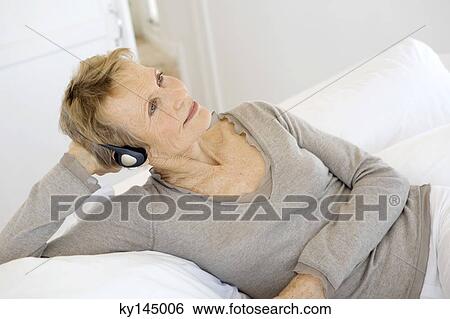 Stock Photograph - Senior woman lying on bed, listening to music with headphones. Fotosearch