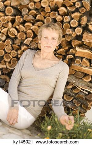 Stock Image - Senior woman sitting by woodpile. Fotosearch