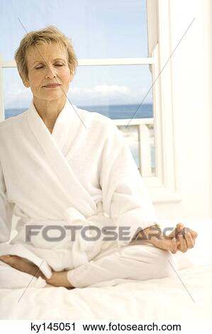 Stock Image - Senior woman sitting cross-legged on bed. Fotosearch