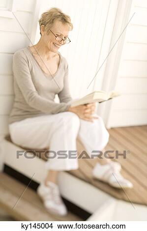 Smiling senior woman reading a book, sitting on wooden terrace View Large Photo Image Picture - Smiling senior woman reading a book, sitting on wooden terrace. Fotosearch