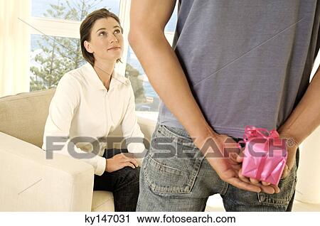 Man hiding gift behind his back, facing young woman View Large Photo Image Stock Image - Man hiding gift behind his back, facing young woman. Fotosearch