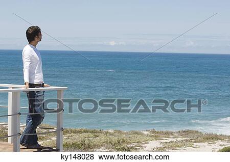 Stock Photo - Young man leaning against balustrade, looking out to sea. Fotosearch