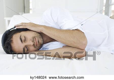 Picture - Young man listening to music with headphones, lying on bed. Fotosearch