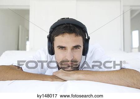Young man lying on bed, listening to music with headphones View Large Photo Image Stock Photo - Young man lying on bed, listening to music with headphones. Fotosearch
