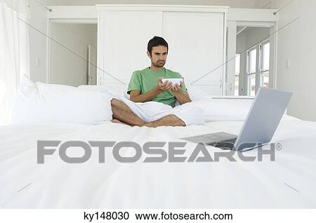 Young man sitting cross-legged on a bed, looking at laptop View Large Photo Image Stock Image - Young man sitting cross-legged on a bed, looking at laptop. Fotosearch