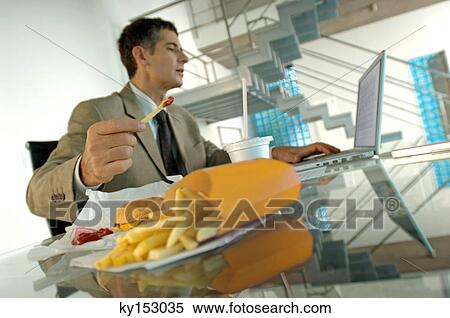 Mid adult businessman eating French fries, using laptop View Large Photo Image Stock Photography - Mid adult businessman eating French fries, using laptop. Fotosearch