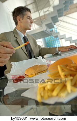 Stock Image - Mid adult businessman having burger and French fries, using laptop. Fotosearch