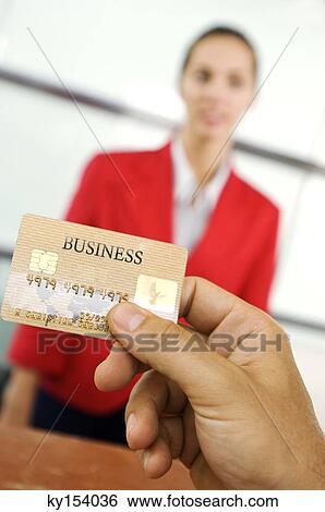 Stock Photograph - Human hand holding credit card with businesswoman standing in background, close-up. Fotosearch