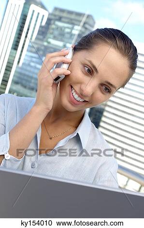Stock Image - Young businesswoman using laptop and mobile phone, smiling. Fotosearch