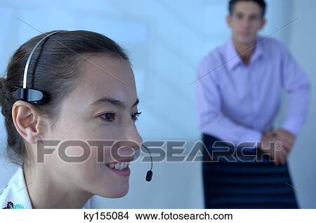 Businesswoman using headset with colleague standing in background View Large Photo Image Picture - Businesswoman using headset with colleague standing in background. Fotosearch