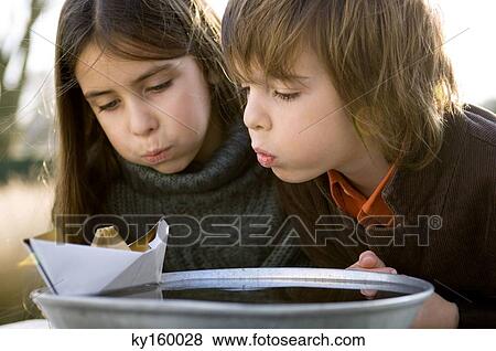 Stock Photo - Close-up of two children blowing the paper boat on a bowl of water. Fotosearch