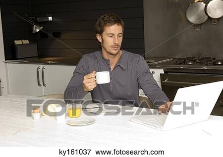 Stock Photo - Mid adult man working on a laptop and having breakfast in the kitchen. Fotosearch