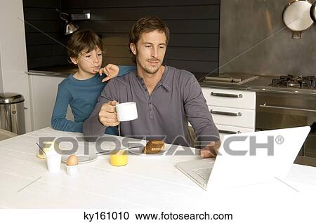 Stock Image - Mid adult man working on a laptop with his son standing behind him. Fotosearch