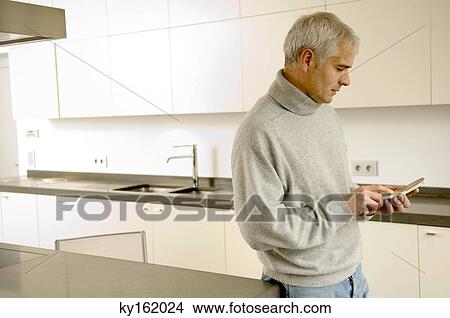 Mature man standing in the kitchen and text messaging on a mobile phone View Large Photo Image Picture - Mature man standing in the kitchen and text messaging on a mobile phone. Fotosearch