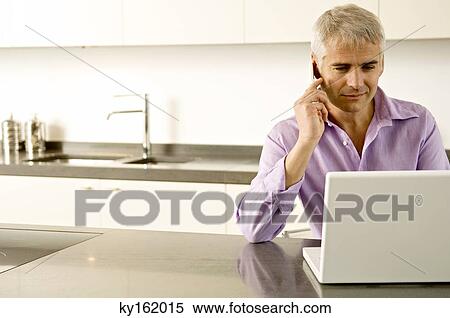 Mature man working on a laptop while talking on a mobile phone View Large Photo Image Stock Photography - Mature man working on a laptop while talking on a mobile phone. Fotosearch