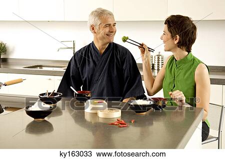 Stock Image - Mid adult woman feeding food to a mature man in the kitchen. Fotosearch