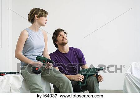 Stock Photo - Mid adult man and a young woman sitting in a room and holding drills. Fotosearch