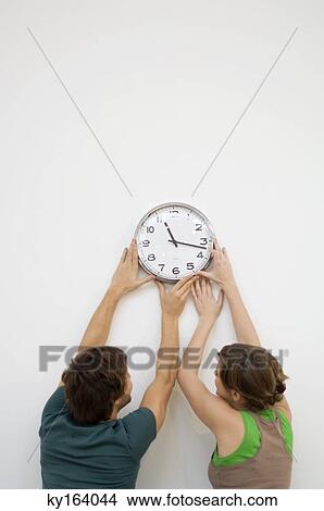 Picture - Rear view of a mid adult man and a young woman mounting a clock on the wall. Fotosearch