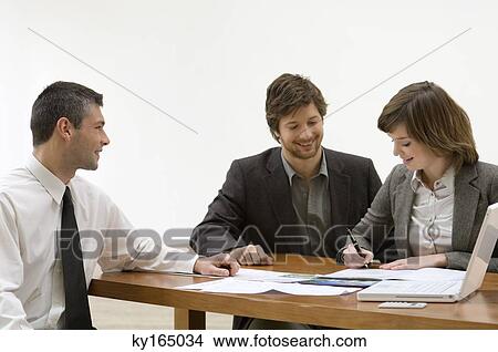 Young woman filling a document with a mid adult man and a real estate agent sitting with her View Large Photo Image Picture - Young woman filling a document with a mid adult man and a real estate agent sitting with her. Fotosearch