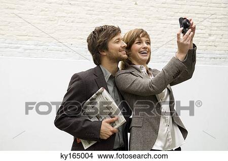 Picture - Young woman taking a picture with a digital camera and a mid adult man standing behind her. Fotosearch