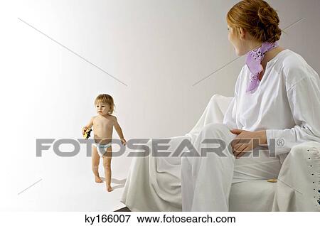 Young woman sitting on a couch and looking at her son walking with a toy car View Large Photo Image Stock Photo - Young woman sitting on a couch and looking at her son walking with a toy car. Fotosearch