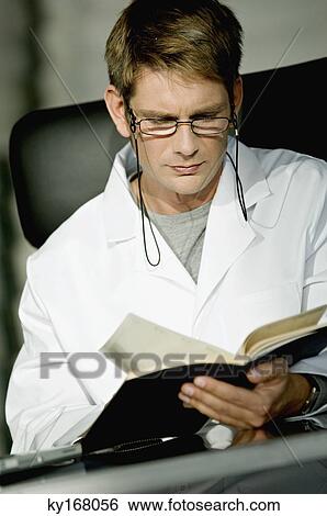 Close-up of a male doctor sitting at a desk and reading a notebook View Large Photo Image Stock Photograph - Close-up of a male doctor sitting at a desk and reading a notebook. Fotosearch