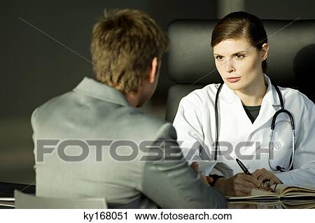 Stock Image - Female doctor sitting at a desk with a male patient in her office. Fotosearch